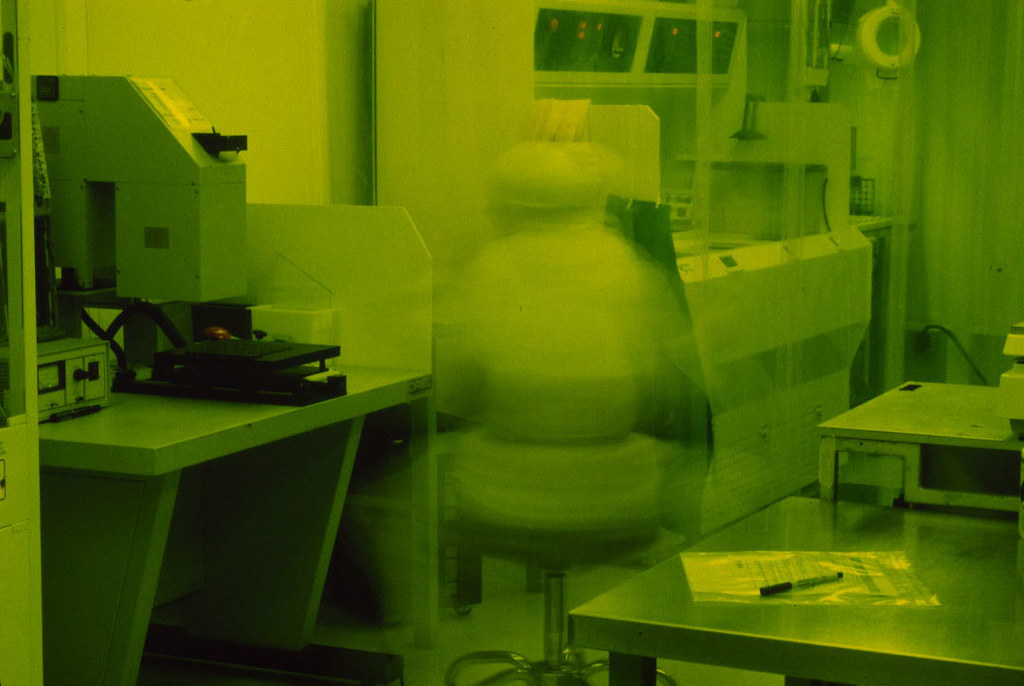 A cleanroom worker in a white bunny suit, blurred in long exposure among lithography tools