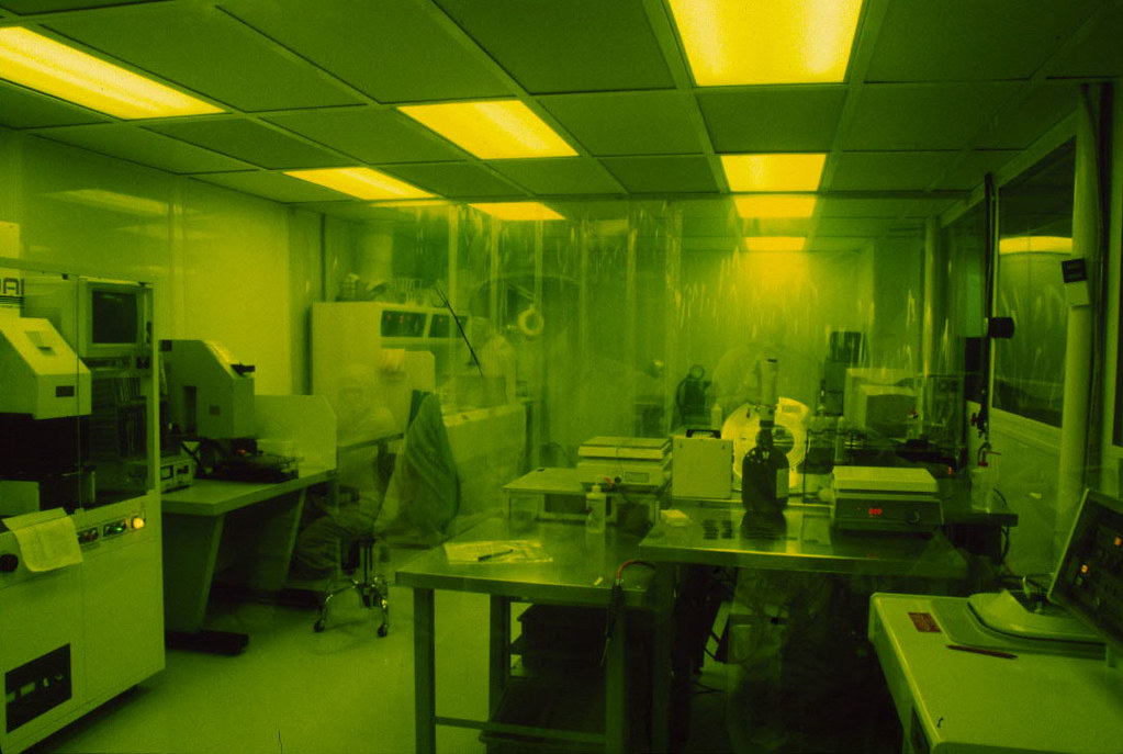 A photolithography cleanroom glowing under yellow sodium light; a technician in a full cleanroom suit works at a bench