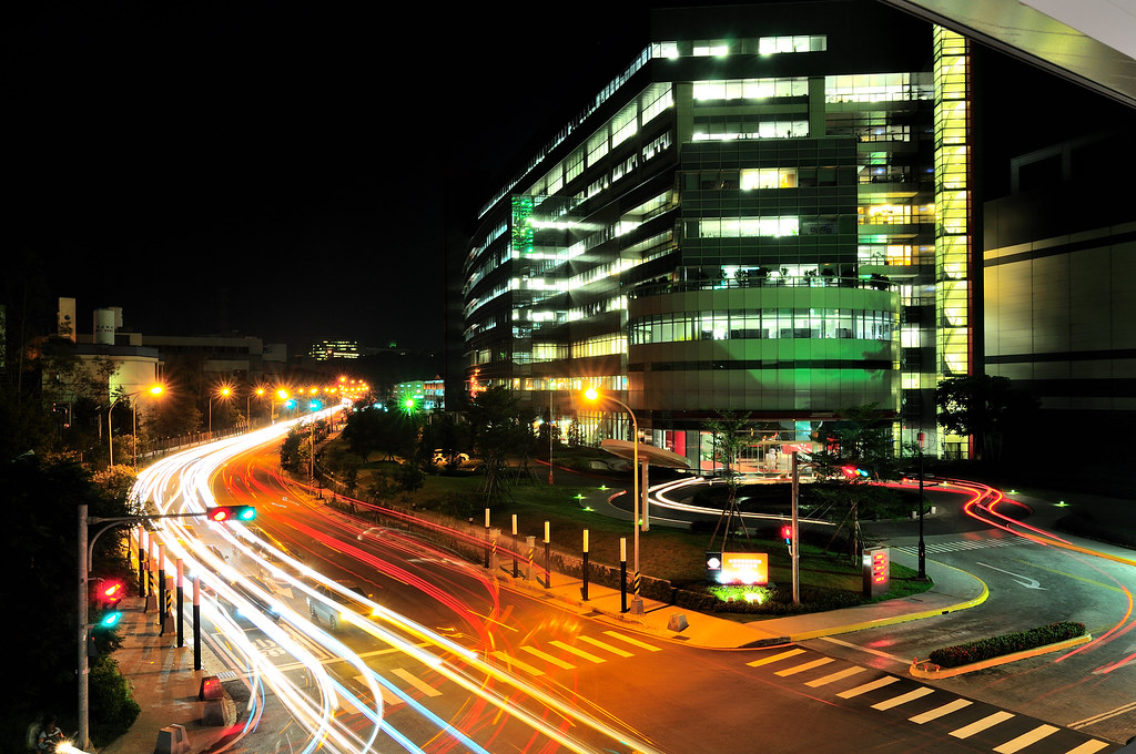 TSMC Fab 12 in Hsinchu at night, windows glowing, car headlights drawing long light trails around the building